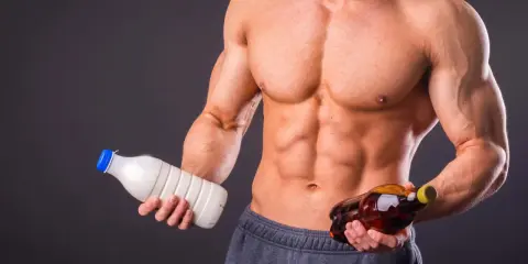 Muskulöser Mann hält Milch und eine Flasche mit bernsteinfarbener Flüssigkeit. Die Frage: Alkohol und Muskelaufbau – eine gute Kombination?