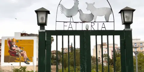 Cancello verde con insegna "FATTORIA" e sagome di animali. A sinistra, murale di un uomo su trono. Danisinni, Palermo.
