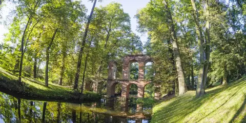 Kamienne ruiny mostu w Arkadii Nieborów, otoczone bujną zielenią i odbijające się w spokojnej wodzie.
