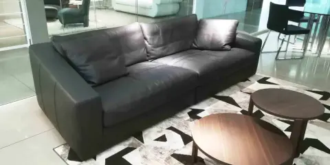 Un elegante divano angolare in pelle nera, con tavolini rotondi in legno, su un tappeto geometrico. Un frighetto è visibile in lontananza.