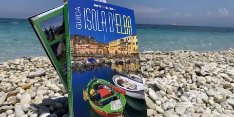 Guida turistica "Isola d'Elba" su una spiaggia di ciottoli, con il mare azzurro sullo sfondo.