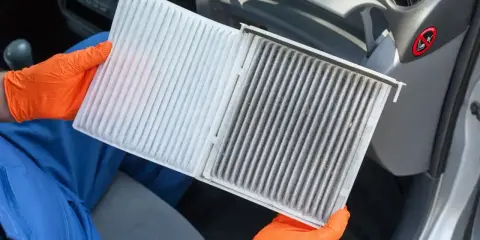 Ręce w pomarańczowych rękawiczkach pokazują brudny filtr kabinowy Toyoty Yaris, gotowy do wymiany.