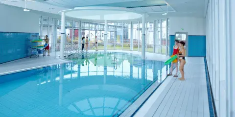 Menschen in Badekleidung am Beckenrand eines Hallenbades mit türkisfarbenem Wasser.