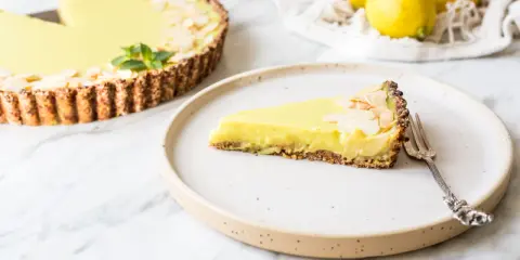 Ein Stück zitronentarte vegan mit Mandelsplittern auf einem Teller. Daneben eine ganze Tarte und frische Zitronen.