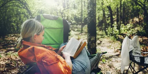 Dziewczyna z blond warkoczem czyta książkę, siedząc na krześle turystycznym podczas biwaku w lesie. Słońce przebija się przez liście drzew.