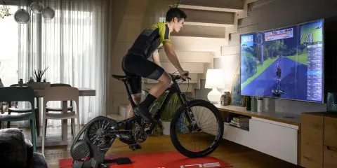 Mężczyzna ćwiczy na rowerze stacjonarnym, korzystając z aplikacji do trenażera, która wyświetla wirtualną trasę na dużym ekranie telewizora.
