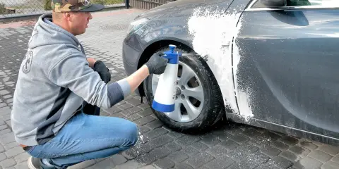 Mężczyzna w czapce z daszkiem i jeansach kuca przy samochodzie, rozpylając pianę na błotnik. Czy można naprawiać auto pod blokiem? Tak, jeśli to drobna naprawa lakieru.