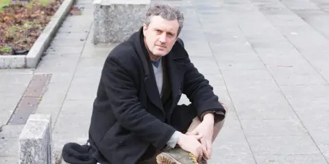 Uomo con cappotto nero seduto all'aperto, ricorda le atmosfere dei rocco schiavone libri.