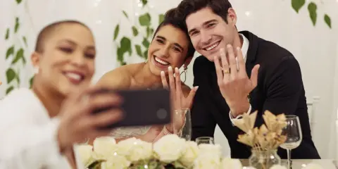 Brautpaar zeigt Ringe für ein Hochzeitsfoto, während eine Freundin ein Selfie macht.
