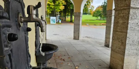 Erfrischendes Wasser fließt aus einem historischen Brunnen im Kurpark Bad Hermannsborn. Herbstlaub liegt auf dem gepflasterten Weg.