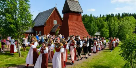 En procession av musiker i folkdräkter, som spelar fiol och gitarr, går förbi en röd träkyrka. Många människor i traditionella kläder, vilka firar midsommar, följer efter.