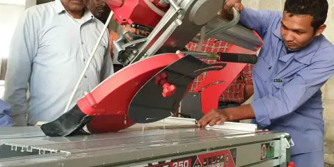 Un trabajador realiza un corte a inglete preciso en un perfil metálico con una cortadora de azulejos, mientras otros observan.