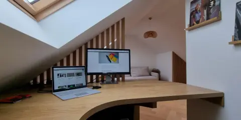 Drewniane biurko na poddaszu pod skosem, z laptopem i monitorem. W tle kanapa i płyty winylowe na ścianie.