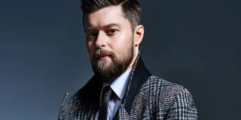Mężczyzna z brodą i starannie ułożonymi włosami, w eleganckim płaszczu w kratę. Remigiusz Mróz wiek nie jest widoczny, ale styl sugeruje dojrzałość.