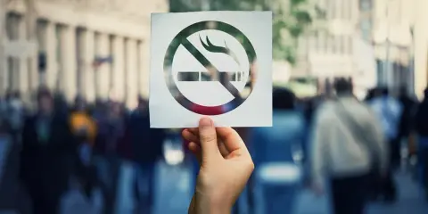Dłoń trzyma kartkę z symbolem zakazu palenia papierosów na tle tłumu ludzi.