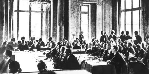 Männer in Anzügen sitzen an einem langen Tisch in einem prunkvollen Saal. Sie diskutieren die Bedingungen des Versailler Vertrags.