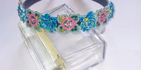 Elegancka opaska z kamieniami w kształcie kwiatów, ozdobiona niebieskimi różami i różowymi detalami, błyszczy na szklanym flakonie.