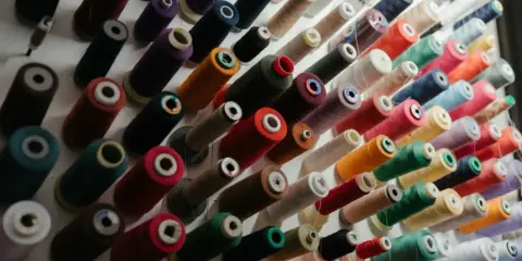 Mnóstwo kolorowych nici do szycia ułożonych na półce. Różne rodzaje nici do szycia, od cienkich po grube, w odcieniach zieleni, czerwieni, fioletu i wielu innych.