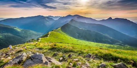 Widok na pasmo górskie o zachodzie słońca. Na pierwszym planie kamienie i trawa, w oddali zielone zbocza i skaliste szczyty. Kasprowy Wierch wysokość robi wrażenie.