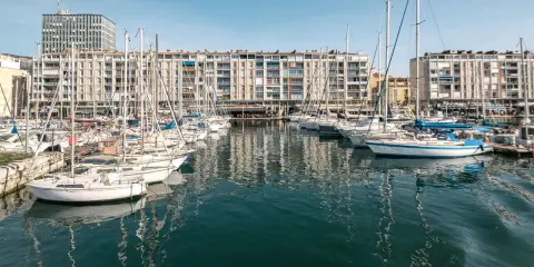 Barche a vela ormeggiate nel porto di Tolone, con edifici residenziali sullo sfondo e il cielo azzurro.