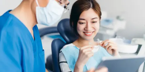 Uśmiechnięta pacjentka trzyma przezroczyste nakładki, a dentysta w maseczce i czepku pokazuje jej coś na tablecie. To pokazuje, jak zwracać się do dentysty.