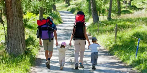 Familia con mochilas camina por un sendero boscoso, ideal para rutas para hacer con niños.