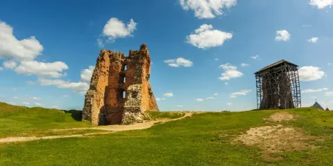 Ruiny ceglanego, opuszczonego zamku na zielonym wzgórzu pod błękitnym niebem z chmurami. Obok wieża w rusztowaniach.