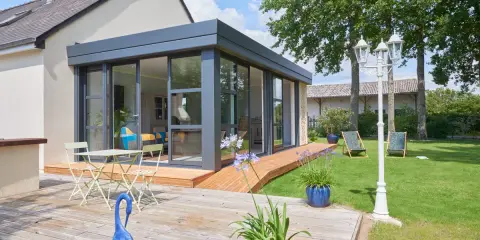 Extension toit plat sur maison ancienne, avec terrasse en bois, mobilier de jardin et vue sur un jardin verdoyant.