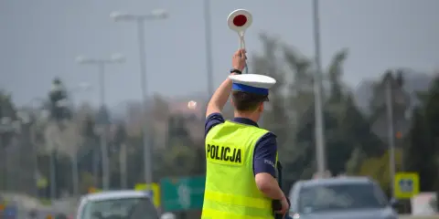 Policjant kieruje ruchem, podnosząc znak stop. W tle widać samochody i słupy oświetleniowe, sugerujące podwyższone **stopnie alarmowe**.