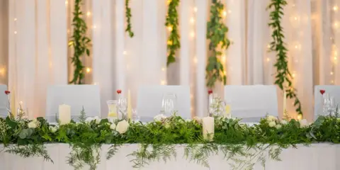Stół weselny w stylu greenery, udekorowany bujną zielenią, białymi różami i świecami. Tło tworzą zwiewne zasłony i delikatne światełka.