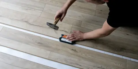 Mann verlegt Laminat mit Hammer und Schlagklotz. Die neuen Dielen werden präzise zusammengefügt.