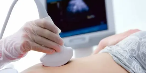 Ręka w rękawiczce trzyma sondę USG, która bada brzuch. Na ekranie widać obraz z badania, być może to test pappa.