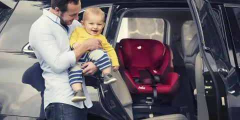 Tata z uśmiechniętym dzieckiem przy samochodzie, obok czerwonego fotelika Joie i-Spin 360.