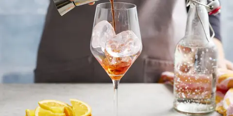 Ein Aperol-Spritz-Cocktail mit Prosecco wird zubereitet. Eiswürfel im Glas, daneben Orangenstücke und eine Flasche Sprudelwasser.