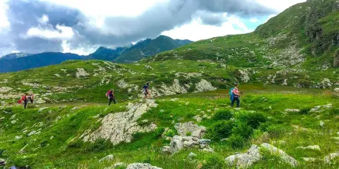 Escursionisti attraversano un paesaggio verdeggiante in una località montagna Piemonte, con rocce e nuvole minacciose all'orizzonte.