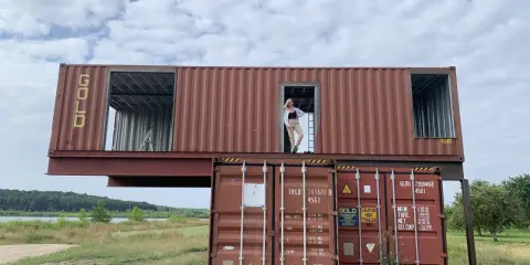Dom z kontenerów w budowie. Kobieta stoi w otwartych drzwiach górnego kontenera, patrząc na krajobraz.