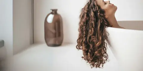 Une femme aux cheveux bouclés se détend dans une baignoire. Elle se demande combien de shampoing par lavage est idéal pour ses cheveux.