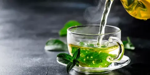 Un thé vert vif est versé dans une tasse transparente, infusant des feuilles de menthe poivrée. La vapeur s'élève, promettant une boisson rafraîchissante.