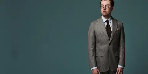 A man in a pinstripe suit, with a brown tie and glasses, stands against a teal background. The jacket length appears to be ideal, hitting just below the seat.
