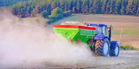 Traktor rozsiewa wapno do ziemi, tworząc chmurę pyłu na polu. W tle las i wzgórza.