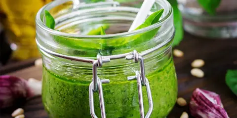 Un pot de pesto ail des ours, vert vif, avec une cuillère blanche, des pignons de pin et de l'ail sur un fond en bois.
