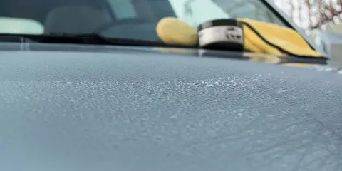 Krople wody na masce samochodu, obok słoiczek z woskiem i żółta mikrofibra. Przygotowanie do woskowania samochodu.