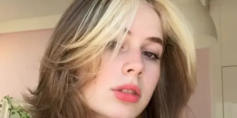 Dziewczyna z burzą blond i brązowych włosów w stylu curly wolf cut, z pasemkami przy twarzy.