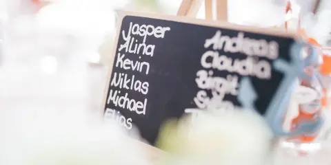 Tablica z listą gości weselnych, na której widnieją angielskie imiona i nazwiska, np. Jasper, Alina, Kevin, Andreas, Claudia.