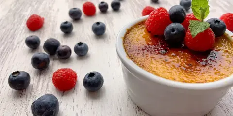 Creme brulee mit karamellisierter Kruste, garniert mit frischen Himbeeren und Blaubeeren, umgeben von weiteren Beeren auf hellem Holz.
