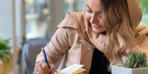 Kobieta w beżowym płaszczu pisze w notesie, tworząc swój własny sonet. Obok leży telefon i sukulent.