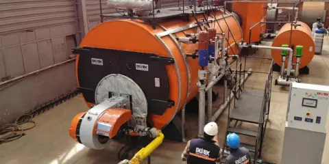 Dos técnicos observan varios tipos de calderas industriales naranjas y grises en una fábrica.