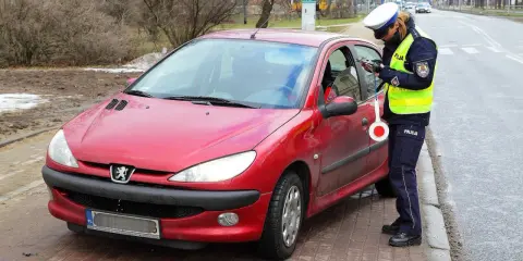 Policjantka w kamizelce odblaskowej i czapce spisuje kierowcę przy czerwonym samochodzie.