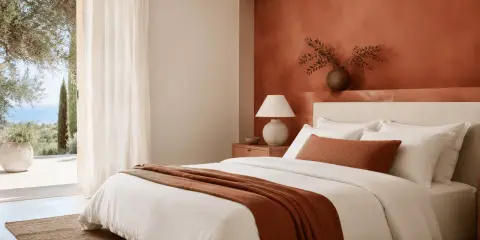 Chambre lumineuse avec mur couleur terracotta, lit blanc, jeté assorti et vue sur la mer.