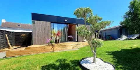 Un charmant pavillon maison moderne avec terrasse en bois, entouré d'un jardin verdoyant et d'un petit olivier.